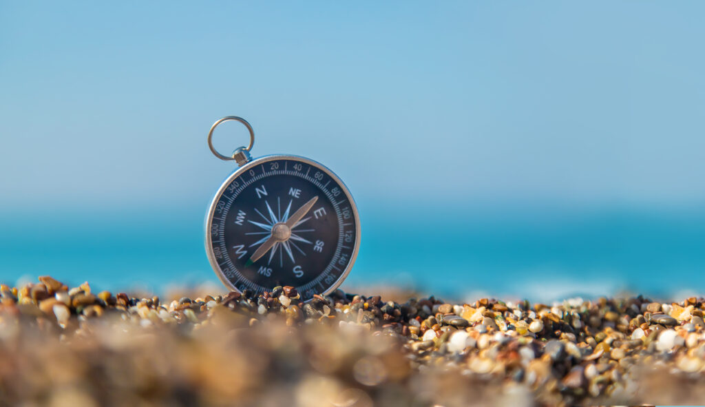 Schwarzer Kompass im Kies am Strand mit Blick aufs Meer – Symbol für neue Ausrichtung und Klarheit im Karriere Coaching.